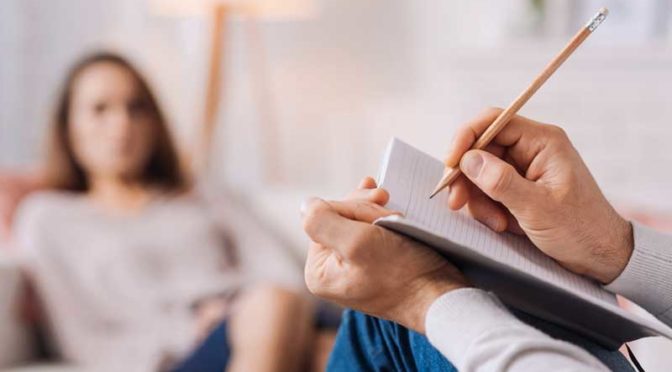 A psychiatrist with a notepad and pencil sitting opposite a woman sitting in a chair